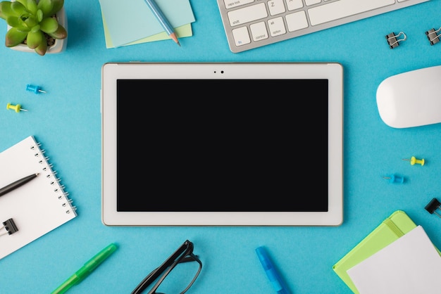 Top view photo of workplace with chancellery keyboard mouse glasses plant and tablet computer with copyspace in the middle on isolated blue background