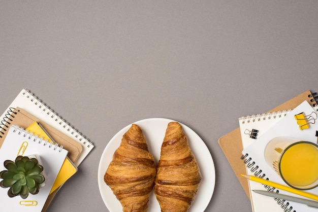 Top view photo of workstation stationery stacks of notepads binder clips pencil pen plant glass of orange juice and plate with croissants on isolated grey background with copyspace