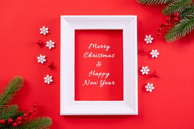 Top view of picture frame, snowflakes, red berries and Christmas tree decoration on red background with text.