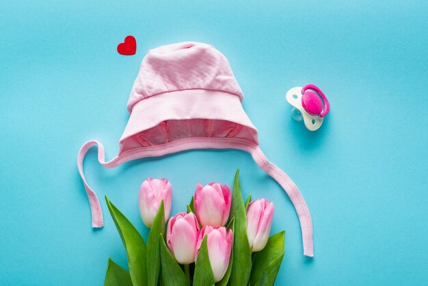 Top view of pink baby hat and tulips near pacifier on blue background concept of mothers day