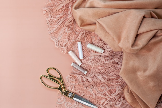 Top view of pink coral coloured lace and silk fabrics with thread spools, calculator on notepad, tape measure, tailoring scissors near cup of coffee on pink background. Evening gowns sewing concept.