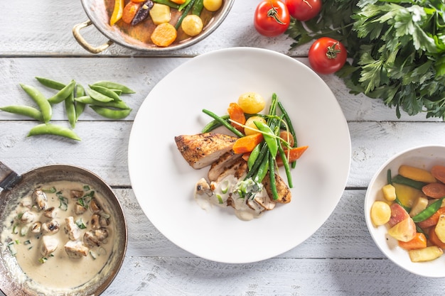 Top view of a plate with cooked vegetables and potatoes with chicken breasts and mushroom sauce in separate bowls.