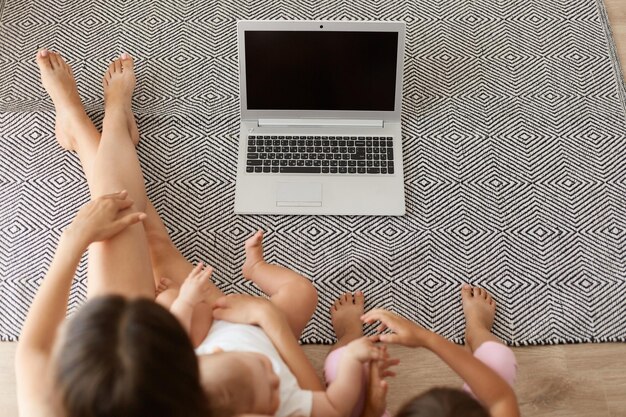 Vista dall'alto ritratto di mamma e figlie seduti sul pavimento del soggiorno e guardare cartoni animati o video divertenti tramite laptop, notebook con schermo vuoto per la tua pubblicità.