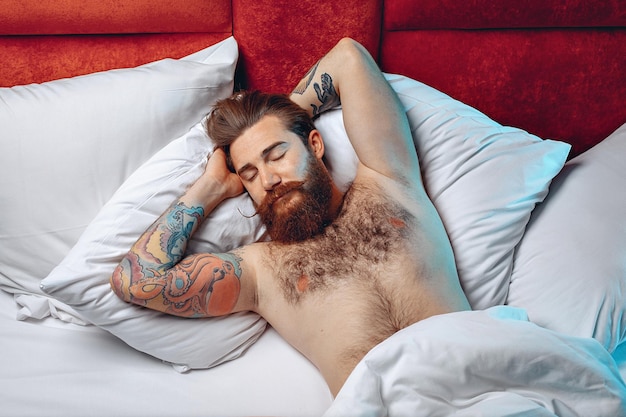 Вид сверху портрет спящего красивого обонятельного мужчины с бородой, усами и татуировкой, лежащего на спине на белой кровати и спящего. Концепция сна