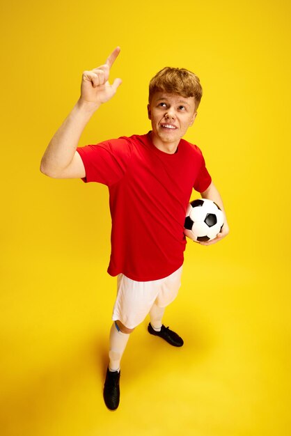 Top view portrait of young guy with freckles in football uniform holding soccer ball against yellow