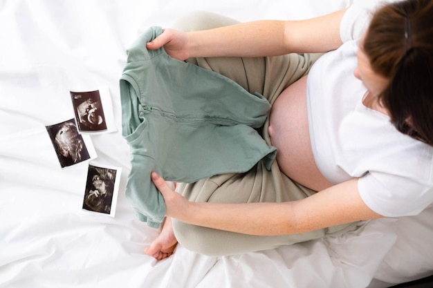 Foto vista dall'alto madre incinta che tiene i vestiti del bambino in mano felice madre in attesa bambino preparazione per il parto ragazza grande pancia avanzata gravidanza concetto di maternità