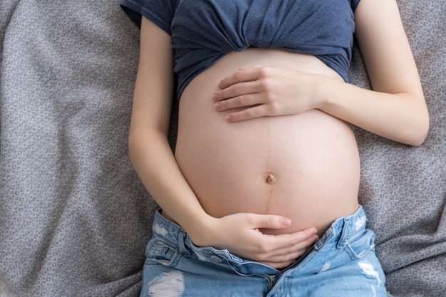 Top view pregnant woman dressed in jeans and t-shirt lying on bed and holding her belly