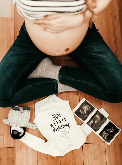 Top view of pregnant woman sitting on the floor. In front of her baby clothes, ultrasound picture and toy.