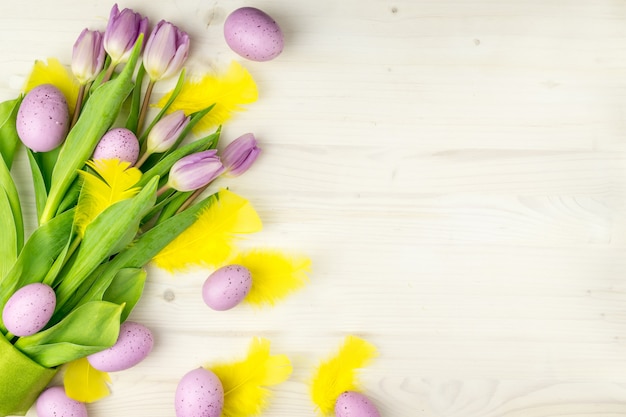 Photo top view of a purple easter eggs with yellow feathers and  purple tulips on a light wood background with message space.