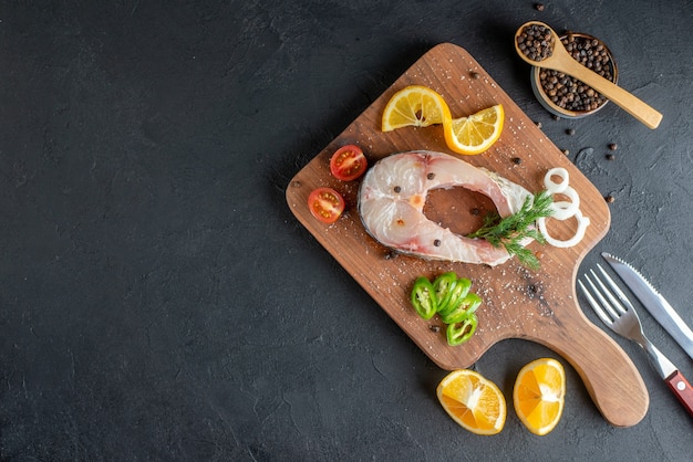 Top view of raw fish and fresh chopped vegetables lemon slices spices on a wooden board cutlery set on black distressed surface