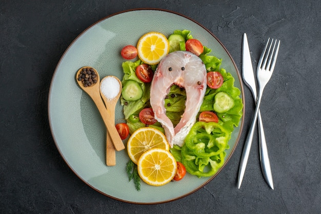 Vista dall'alto di pesce crudo e verdure fresche fette di limone spezie su un piatto grigio e posate su superficie nera