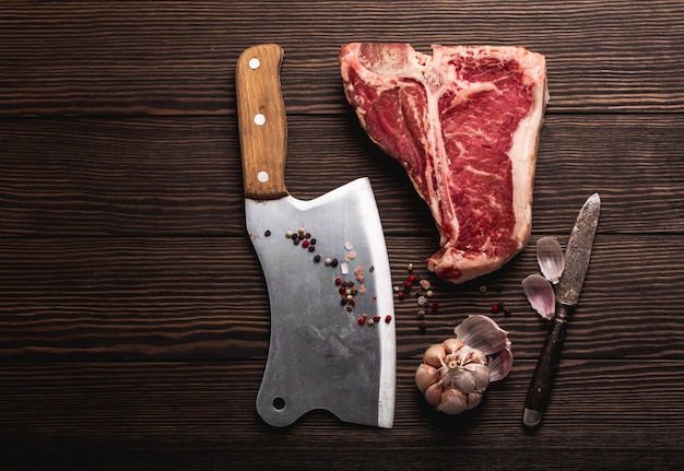 Top view of raw fresh marbled meat steak T-bone, rustic meat cleaver, seasonings on wooden background. Cooking juicy organic steak/butchery concept, healthy clean eating, close up