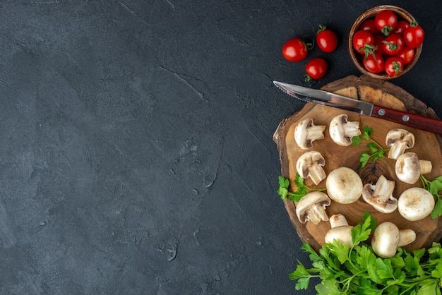나무 판자에 있는 생 버섯과 채소 칼의 맨 위 보기 흰색 수건 신선한 토마토인 토마토는 여유 공간이 있는 검정색 배경 왼쪽에 있는 그릇에 있습니다.