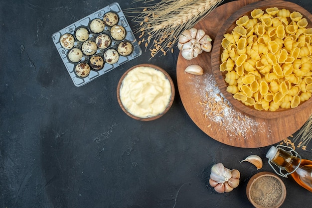 Top view of raw pastas in a brown bowl garlics rice on wooden board flour eggs spikes fallen oil bottle pepper on the left side on black background