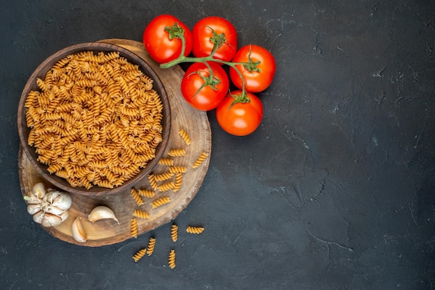 Top view of raw pastas inside and outside brown pot garlics tomatoes on the right side on dark blue color background