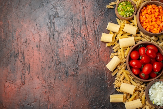 top view raw pastas penne rigatoni spirals chopped vegetables and turmeric in bowls on wooden table with copy place