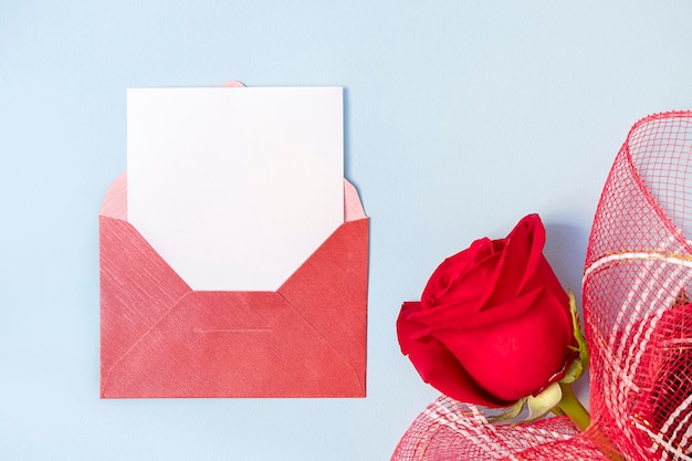 Top view of red envelope and white empty card with beautiful rose on light blue background