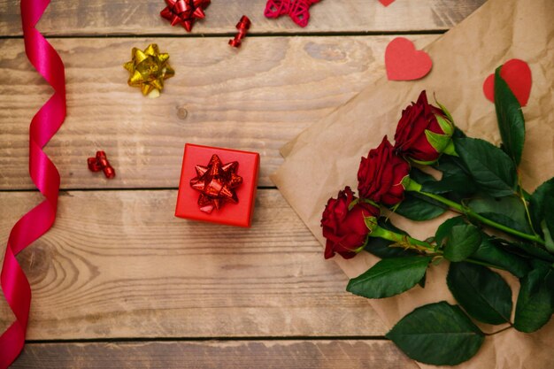 Top view, red gift box. Festive atmosphere, bouquet of red roses and bows on a wooden background.