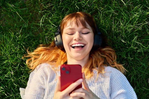 緑の芝生の上のスマート フォンでヘッドフォンで赤髪の若い女性のトップ ビュー