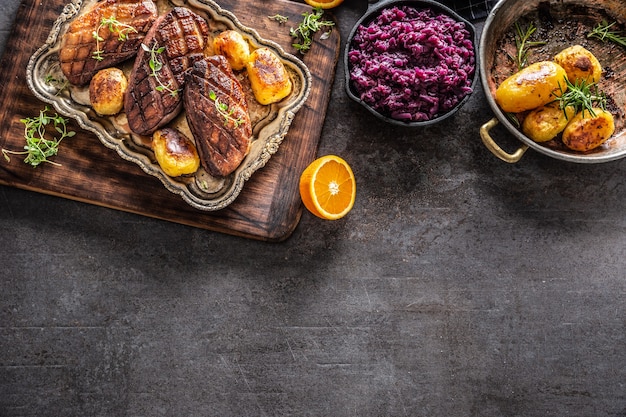 Top view of a roasted duck breasts with oranges, roasted potatoes and red cabbage, on a dark surface.