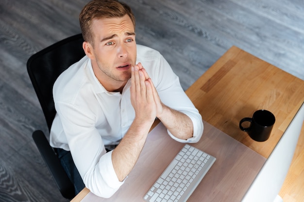 Top view of sad upset young businessman sitting and praying in office