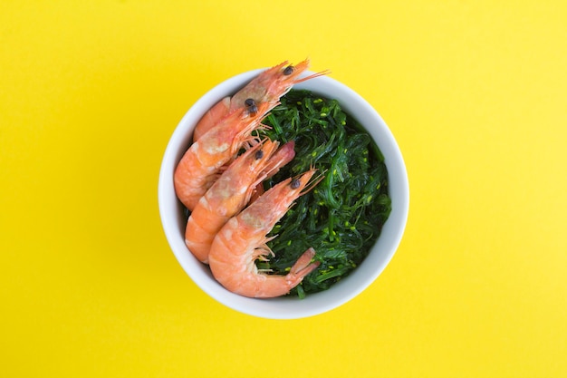 Top view of salad with seaweed and red shrimps in the white bowl on the yellow  background. Close-up.