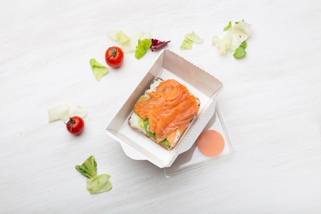 Top view sandwich with soft cheese and red fish lies in the lunch box next to the greens and tomatoes on a white table. Concept of a healthy snack.