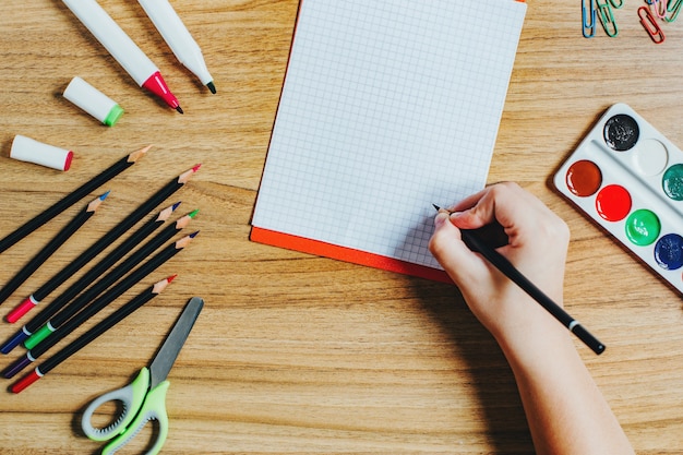 문구 용품, 연필, 펠트 펜, 가위, 페인트, 그리고 공책에 쓰는 어린이의 손이 있는 학생 책상의 상위 뷰
