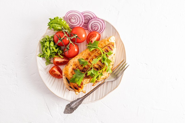 Top view of the serving white plate with juicy grilled chicken fillet with fresh vegetables on a white background a copy of the space