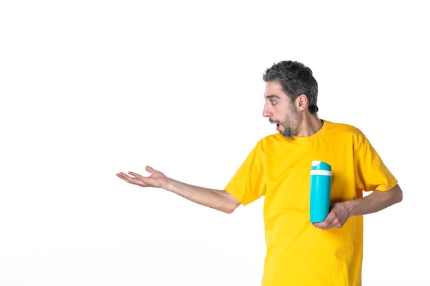 Top view of shocked young male in yellow shirt and showing blue thermos pointing something on the right side on white background