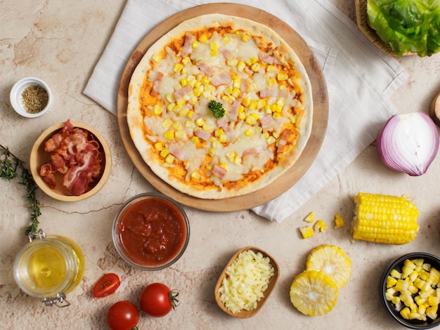 Top view shot of delicious tasty juicy cheesy corn and ham Italian pizza placed on wooden board on party table around with other ingredients sauce olive oil bacon tomatoes cheese and vegetables