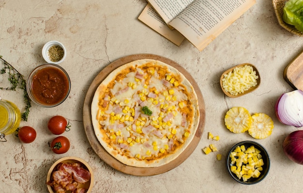 Top view shot of delicious tasty juicy cheesy corn and ham Italian pizza placed on wooden board on party table around with other ingredients sauce olive oil bacon tomatoes cheese and vegetables
