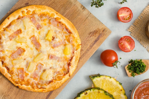 Photo top view shot of delicious tasty juicy of hawaiian traditional italian crust thin crispy ham and pineapple pizza placed on wooden cutting board with ingredients sliced tomatoes ketchup and salt