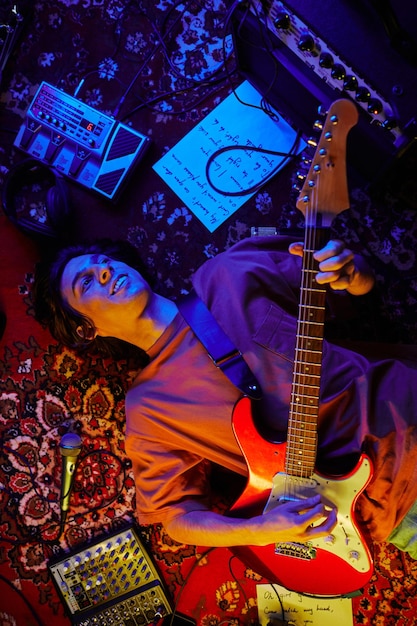 Top view shot of smiling young man playing guitar while lying on carpet in neon light