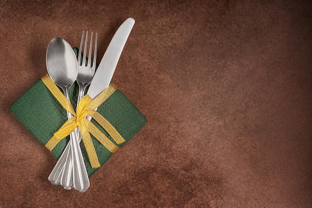 Top view of silver fork, spoon and knife on green napkin with golden band served on brown table