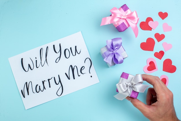 top view small gifts in male hand red and pink hearts will you marry me written on paper on blue background