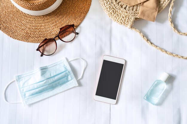 Top view of smartphone, travel accessories, surgical mask and sanitizer gel on bed in hotel room