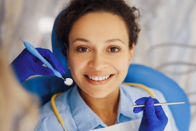 Вид сверху на улыбающуюся женщину, смотрящую в камеру во время стоматологической процедуры, в то время как руки стоматолога используют инструменты