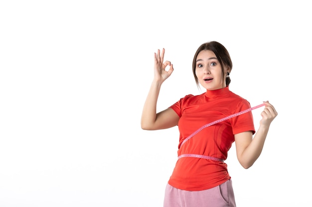 Top view of a smiling young lady gathering her hair dressed in redorange blouse and holding metre measuring her waist making eyeglasses gesture on white background