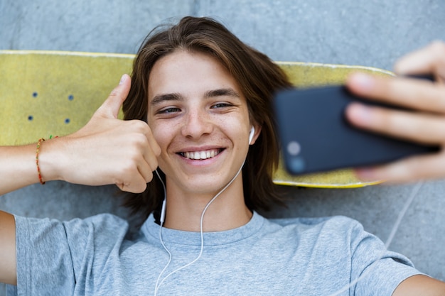 Foto vista dall'alto di un giovane ragazzo adolescente sorridente che trascorre del tempo allo skate park, posa su uno skateboard, prendendo un selfie