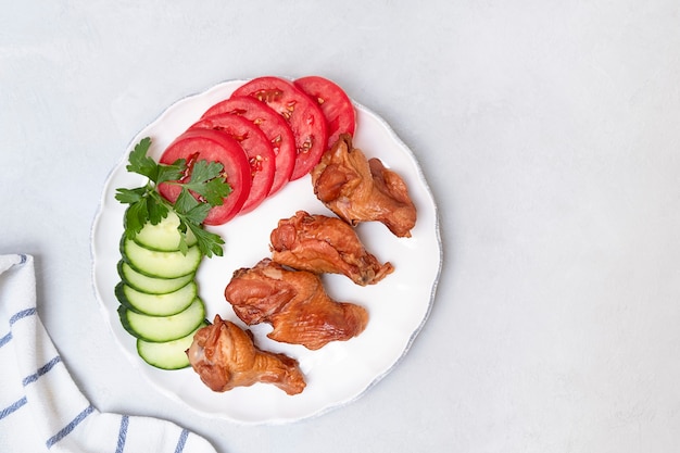 Top view of smoked wings with fresh vegetables on white plate on neutral grey background with space ...