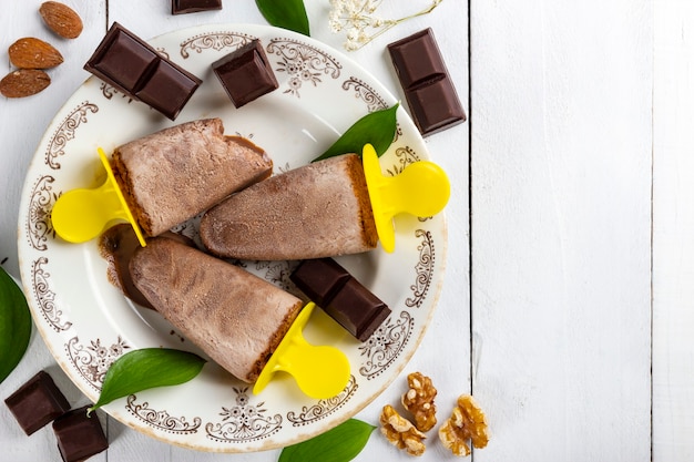 Top view of some delicious and refreshing chocolate ice cream lollies in a vintage plate on a white wooden table covered with pieces of chocolate, nuts, almonds and leaves from nature