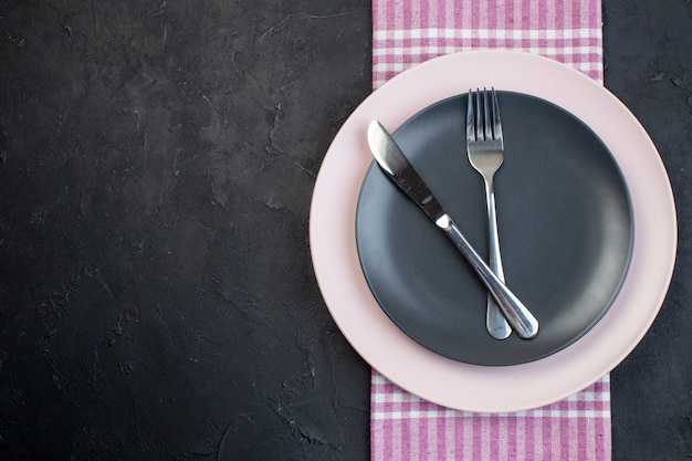 Top view of stainless cutlery set on colorful ceramic empty plates on pink stripped towel on the left side on black background with free space