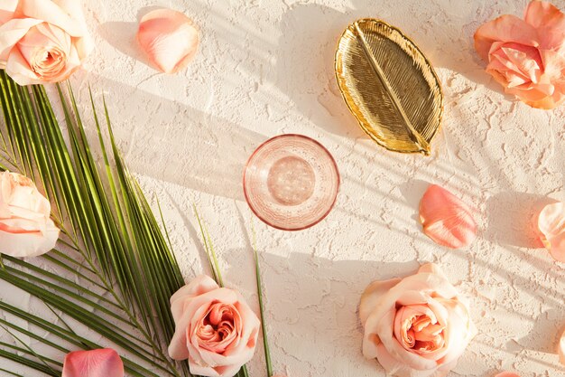 Top view of stylish composition with pink peony roses flowers, tropical palm leaf, golden plate and glass on pastel textured