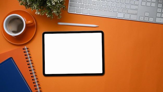 Top view of stylish workspace with digital tablet , notebook, coffee cup and plant on orange background.