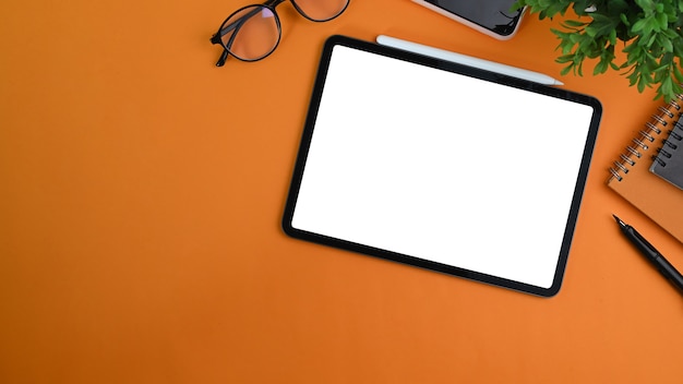 Top view of stylish workspace with digital tablet, notebook, glasses and plant on orange background.