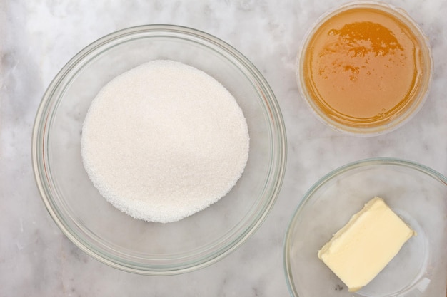 Top view of sugar honey and butter in glass bowl as ingredients of cake medovik on marble background