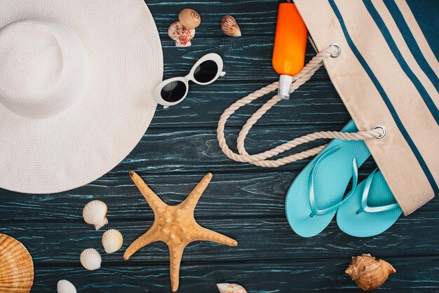 Top view of sun hat near bag flip flops and seashells on dark wooden surface