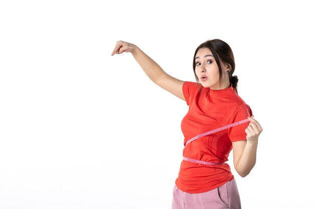 Top view of a surprised young lady gathering her hair dressed in redorange blouse and holding metre measuring her waist pointing down on white background