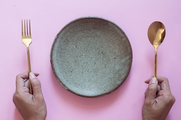 Top view of table, woman hands holding fork and spoon with empty plate on pink background